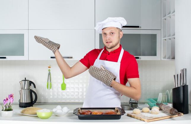 男厨师俯视图：男厨师戴着支架站在桌子后面 上面放着糕点鸡蛋磨碎器 在白色厨房的右侧展示着一些东西帽子立场鸡蛋