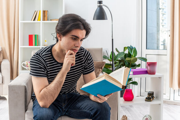 男人穿着休闲服的年轻人脸上带着沉思的表情坐在客厅的椅子上看书沉思阅读表情
