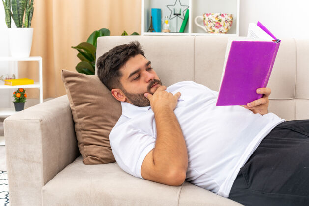 休闲一个穿着休闲服的年轻人 一脸严肃地拿着书在家里度过周末 躺在客厅的沙发上年轻光阅读