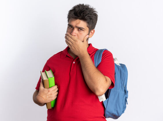 个人年轻的留着胡子的学生 穿着红色马球衫 背着背包 拿着笔记本 手捂着嘴站在白色的墙上背包马球年轻人