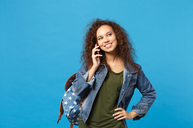 女性年轻的非洲裔美国女孩十几岁的学生在牛仔服装 背包举行电话隔离在蓝色的墙壁上牛仔裤人民族