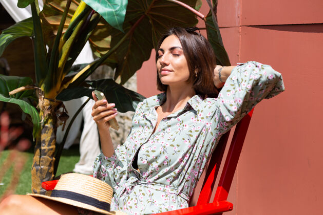 高兴阳光明媚的日子里 穿着夏装的美女坐在后院的椅子上 手里拿着手机欢呼热带花园