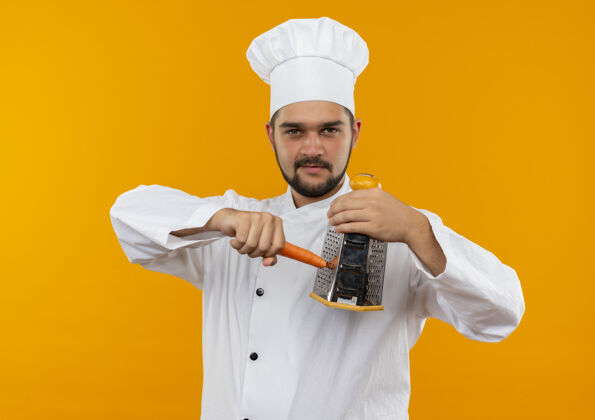 胡萝卜自信的年轻男性厨师身着厨师制服 用隔离在橙色墙壁上的刨子刨胡萝卜烹饪自信感恩