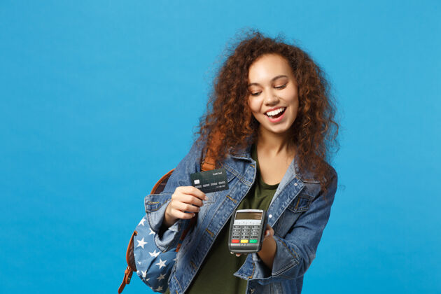 学生年轻的非洲裔美国女孩十几岁的学生在牛仔服装 背包举行信用卡隔离在蓝色的墙上奖励无现金非接触