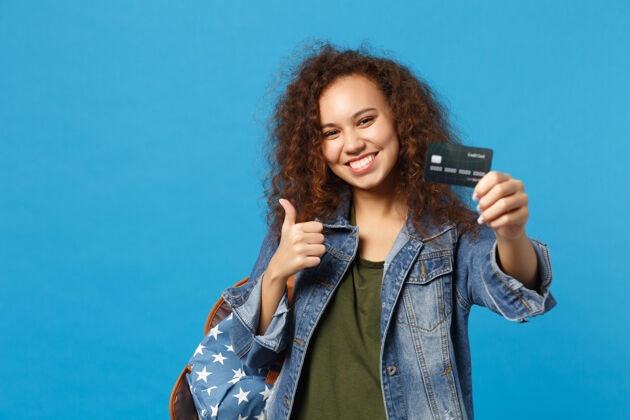 人年轻的非洲裔美国女孩十几岁的学生在牛仔服装 背包举行信用卡隔离在蓝色的墙上欣喜若狂女性奖学金