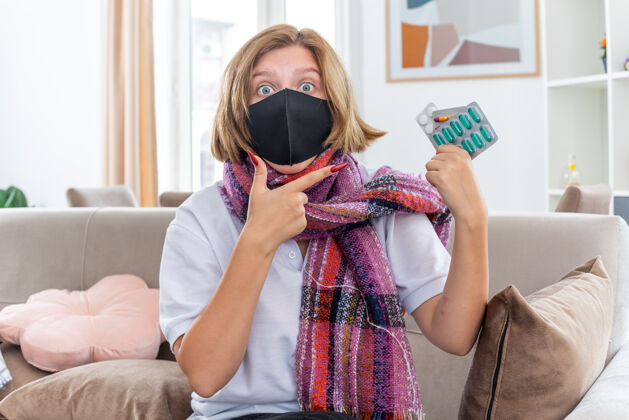感觉不健康的年轻女子 脖子上围着暖和的围巾 戴着面膜 感觉不舒服 生病 患流感 吃感冒药 坐在客厅的沙发上 看起来很困惑流感病困惑