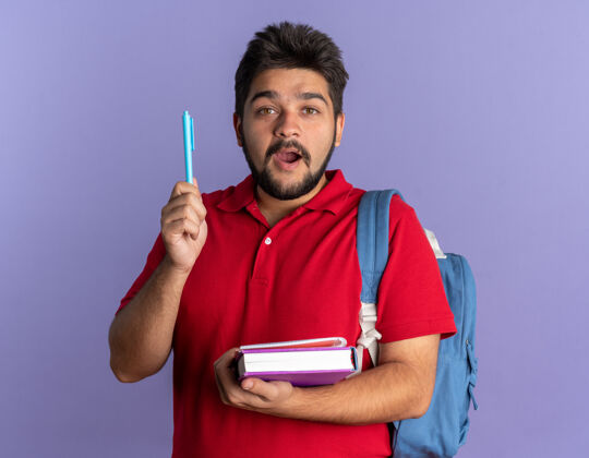 胡须年轻的留着胡子的学生 穿着红色马球衫 背着书包 手里拿着钢笔 站在蓝色的墙上 惊讶地发现自己有了新的想法姿势年轻人惊喜