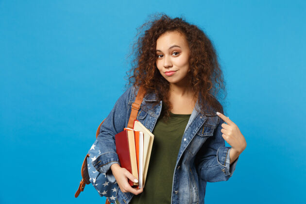 笔记本穿着牛仔服 背着书包的悲伤的年轻学生把书孤立地放在蓝色的墙上青少年非洲女性