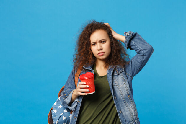 人年轻的非洲裔美国女孩十几岁的学生在牛仔服装 背包举行纸杯隔离在蓝色的墙壁上复制空间马克杯民族