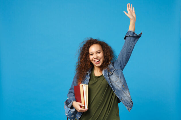 学习穿着牛仔服 背着书包的年轻女学生把书孤立地放在蓝色的墙上胜利毕业成功