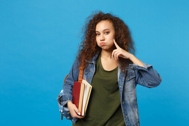 站立穿着牛仔服 背着书包的年轻女学生把书孤立地放在蓝色的墙上手指饮食教育