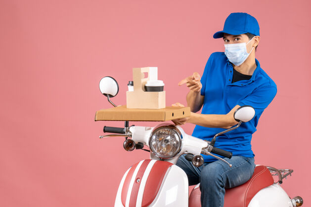 桃顶视图勤劳的快递员戴着医用面罩戴着帽子坐在粉彩桃色背景的踏板车上医学人坐