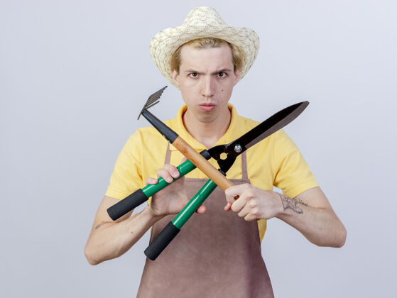 剪刀年轻的园丁 穿着连体衣 戴着帽子 手里拿着小耙子和树篱剪 两手交叉 表情严肃站着花园手