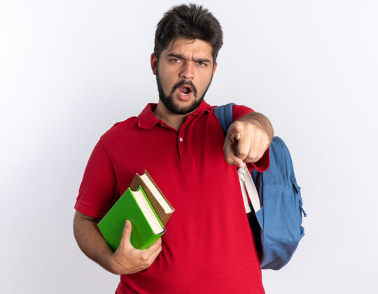 背包年轻的留着胡子的学生 穿着红色马球衫 背着背包 拿着笔记本 用食指指着 愤怒而情绪激动地站在白墙上情绪年轻马球