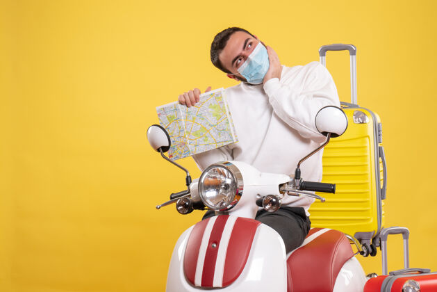 摩托车旅行概念的俯视图 戴着医用面罩的梦幻男孩站在摩托车旁边 黄色手提箱放在上面 手里拿着地图手提箱旅行面具