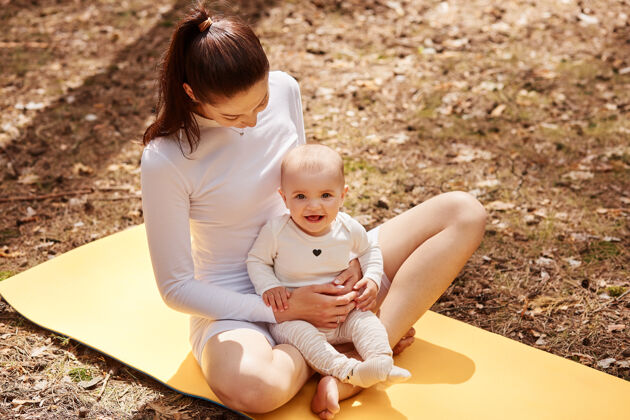 快乐一位年轻的成年女子 深色头发 穿着白色衣服 坐在卡雷马特上 抱着婴儿生活小幼儿
