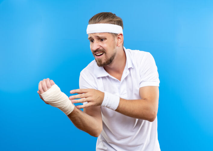 焦虑焦虑的年轻英俊的运动型男子戴着头带和腕带看着他受伤的手腕缠着绷带隔离在蓝色的墙上受伤腕带包