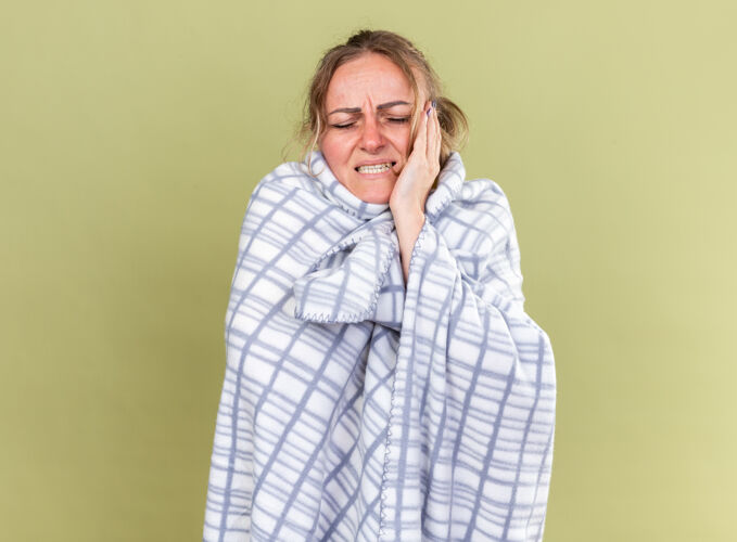 感觉不健康的妇女裹着毯子 站在绿色的墙壁上感觉很可怕 患流感和感冒 头痛得厉害包裹可怕头痛