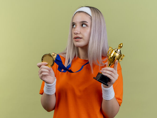 壁板高兴的年轻白人运动女孩戴着背带 戴着头带和腕带 拿着金牌和冠军杯看在一边头带腕带举行