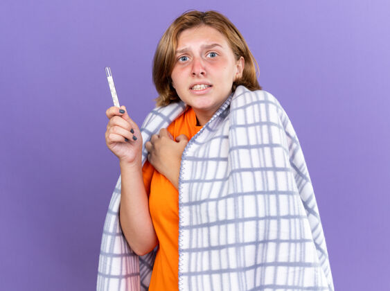 担心不健康的年轻女子裹着温暖的毯子 感觉不舒服 患流感 发烧 用温度计测量体温 站在紫色的墙上 看起来很担心感觉痛苦测量