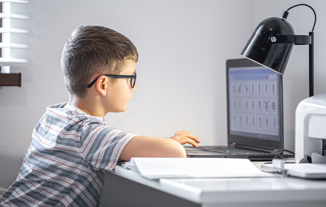 拿着一个戴眼镜的小学生坐在一张桌子旁 手里拿着一台笔记本电脑 在网上做作业书房使用笔记本电脑