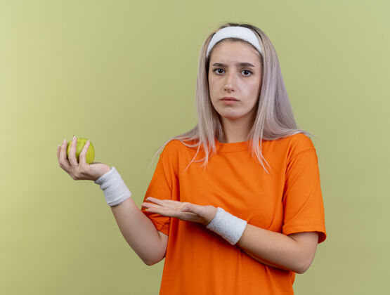 背带无知的年轻白人运动女孩戴着背带 戴着头带和腕带 拿着苹果 指着苹果女孩点举行