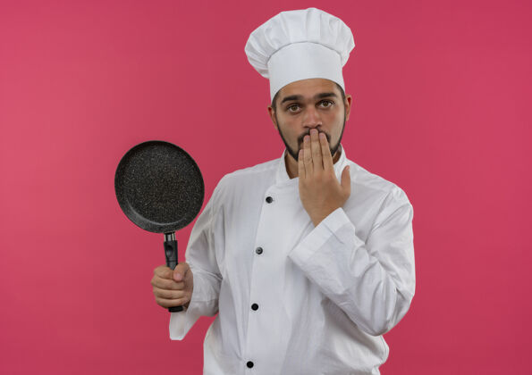 印象穿着厨师制服的年轻男厨师手拿煎锅 把手放在隔离在粉红色墙上的嘴上男性持有平底锅
