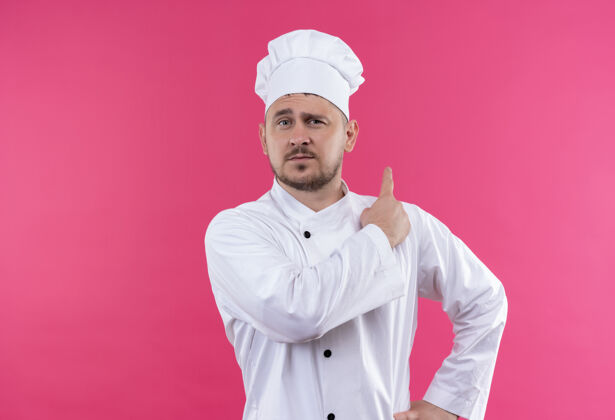 年轻自信的年轻帅哥厨师身着厨师制服指着身后孤零零的粉色墙壁厨师指点自信