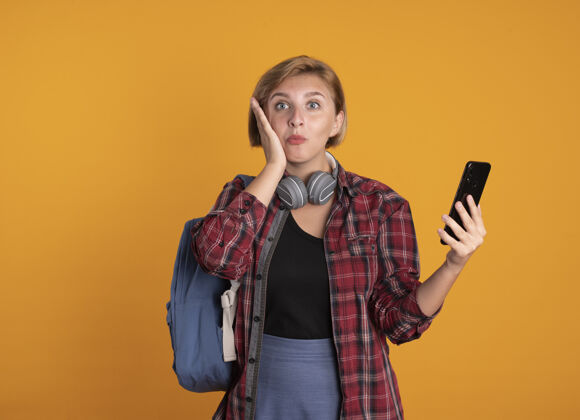 电话惊讶的年轻斯拉夫学生女孩戴着耳机背着背包把手放在脸上拿着电话背包穿橙色