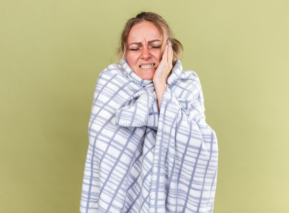 感觉不健康的妇女裹着毯子 站在绿色的墙壁上感觉很可怕 患流感和感冒 头痛得厉害包裹可怕头痛