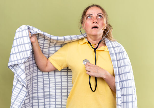 流感不健康的女人裹着毯子 感觉不舒服 患流感和感冒 站在绿色的墙上 用听诊器听心跳 看起来很担心担心不健康包裹