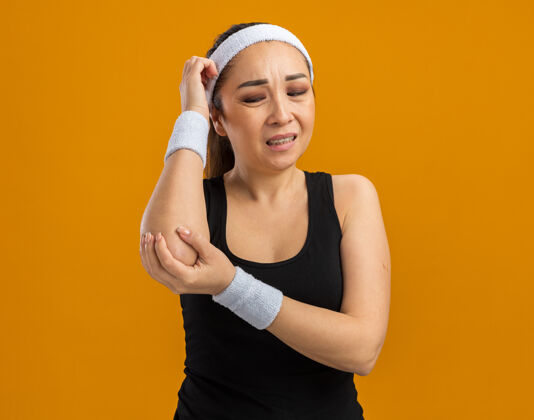 年轻戴着头带和臂章的年轻健身女士站在橙色的墙上 手肘看起来不舒服 感觉疼痛女人健康站立