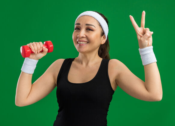 年轻戴着头带和哑铃做练习的年轻健身女士微笑着在绿色墙壁上展示v形标志站立哑铃女子