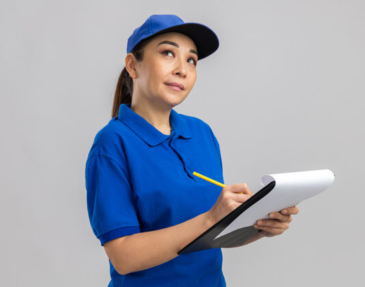 帽子身穿蓝色制服 戴着帽子 拿着写字板和钢笔的年轻女送货员站在白色的墙上 困惑地抬起头来女人钢笔举行