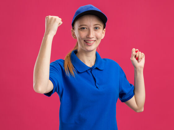 拳头身穿蓝色制服 头戴鸭舌帽的年轻送货员站在粉红色的墙上兴奋而高兴地握紧拳头帽子年轻女孩