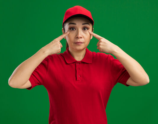 帽子站在绿色的墙上 一位身穿红色制服 头戴鸭舌帽的年轻女送货员用食指指着自己的眼睛 看上去很自信手指女人目录