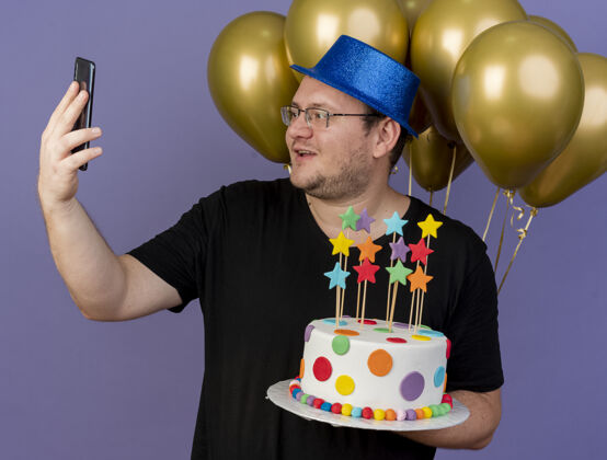 眼镜印象深刻的成年斯拉夫男子戴着眼镜 戴着蓝色派对帽 站在氦气球前 拿着生日蛋糕看着手机光学举行帽子