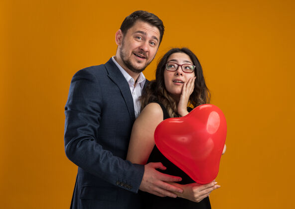 情人节幸福而美丽的情侣们手拿心形的红色气球 在橙色的墙上拥抱庆祝情人节心女人形状
