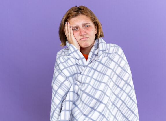 头痛不健康的年轻女子裹着温暖的毯子 感觉不舒服 患流感 发烧 头痛 站在紫色的墙上不健康毛毯温暖