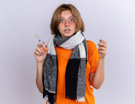 不健康不健康的年轻女子穿着橙色t恤 戴着暖和的围巾 感觉很可怕 患流感 拿着注射器和安瓿站在白色的墙上困惑感情立场迷茫