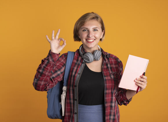 学生微笑着的年轻斯拉夫女学生戴着耳机 背着背包 拿着笔记本和笔 手势ok手势拿着笔记本复制