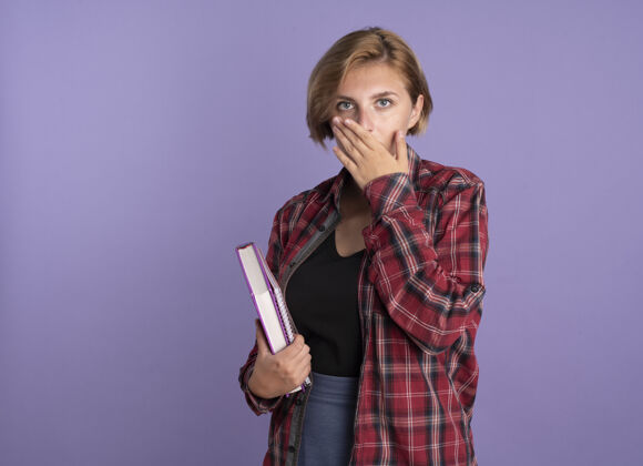 紫色震惊的年轻斯拉夫学生女孩拿着书和笔记本把手放在嘴里持有嘴学生