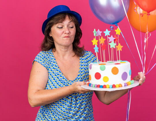 生日快乐快乐的中年妇女戴着派对帽 手里拿着五颜六色的气球 面带微笑地看着生日蛋糕 站在粉色的墙上庆祝生日派对庆祝蛋糕帽子