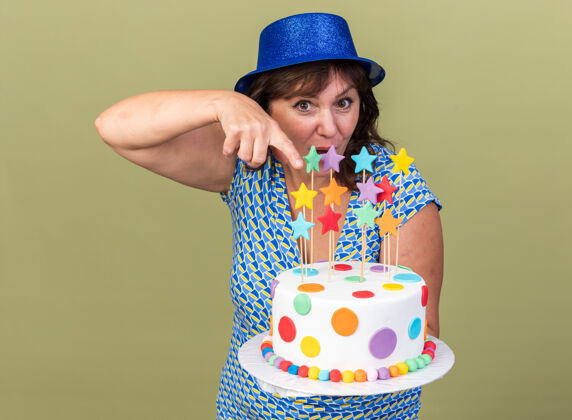 指向中年妇女戴着派对帽 拿着生日蛋糕 用食指指着它 开心又惊喜地站在绿色的墙上庆祝生日派对手指举行生日