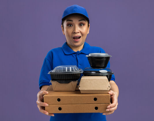 帽子一位身穿蓝色制服 头戴鸭舌帽 手持比萨饼盒和食品包的年轻送货员站在紫色的墙上 她既高兴又惊讶惊喜披萨女人