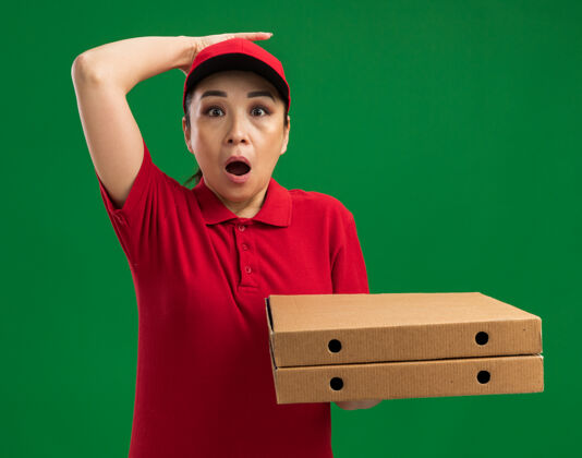 站着年轻的送货员身穿红色制服 戴着帽子 手里拿着披萨盒 手放在头上 站在绿色的墙上 惊讶不已制服年轻女人