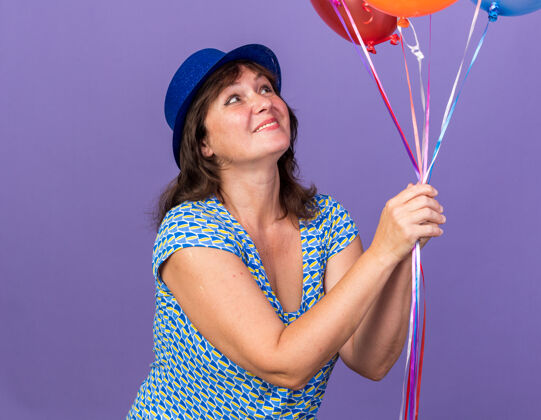 举行快乐快乐的中年妇女 戴着派对帽 手里拿着一束五颜六色的气球 看着它们 微笑着 站在紫色的墙上庆祝生日派对生日束宽