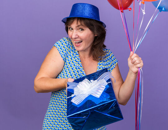 束快乐快乐的中年妇女 戴着派对帽 手持一束五颜六色的气球 站在紫色的墙上 微笑着庆祝生日派对宽生日举行