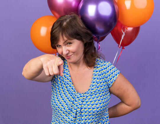 女人快乐的中年妇女 手里拿着一堆五颜六色的气球 食指指着她 微笑着站在紫色的墙上欢庆生日派对束目录气球