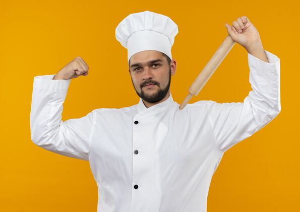 自信身着厨师制服的自信的年轻男厨师把擀面杖放在肩膀上 在橙色的墙上做着强烈的孤立的手势肩膀强壮滚动